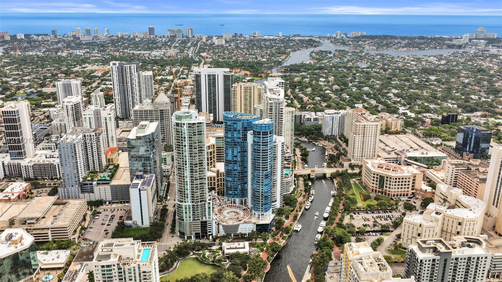 Aerial perspective of urban high-rise development featuring glass-facade towers, proximity to waterway, surrounding metropolitan infrastructure, and coastal horizon