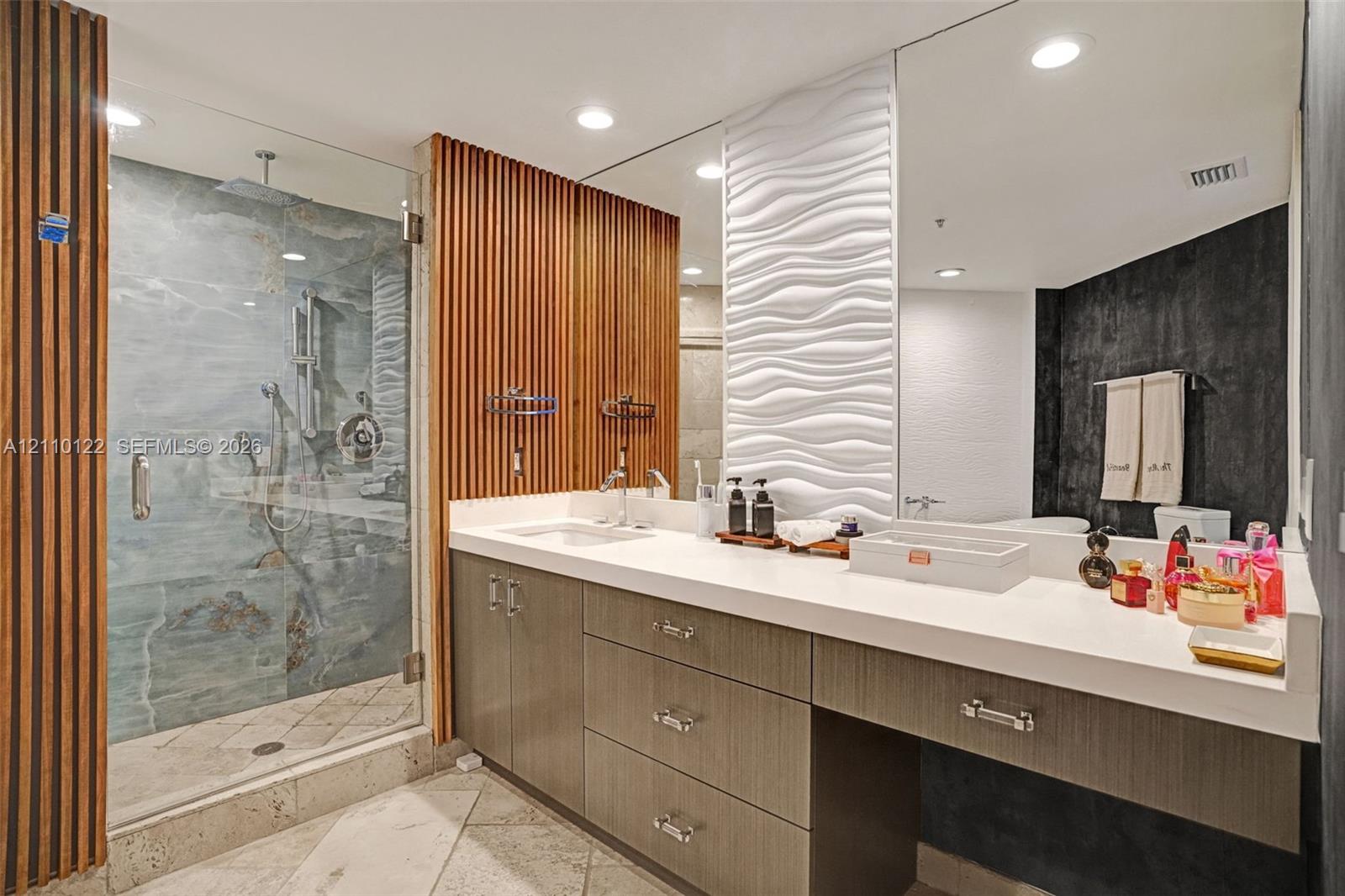 Bathroom featuring a glass-enclosed shower with stone-tile surround, wall-mounted wood-slat paneling, a white dimensional wave-pattern wall accent, a dual-sink vanity with solid-surface countertop, and recessed lighting