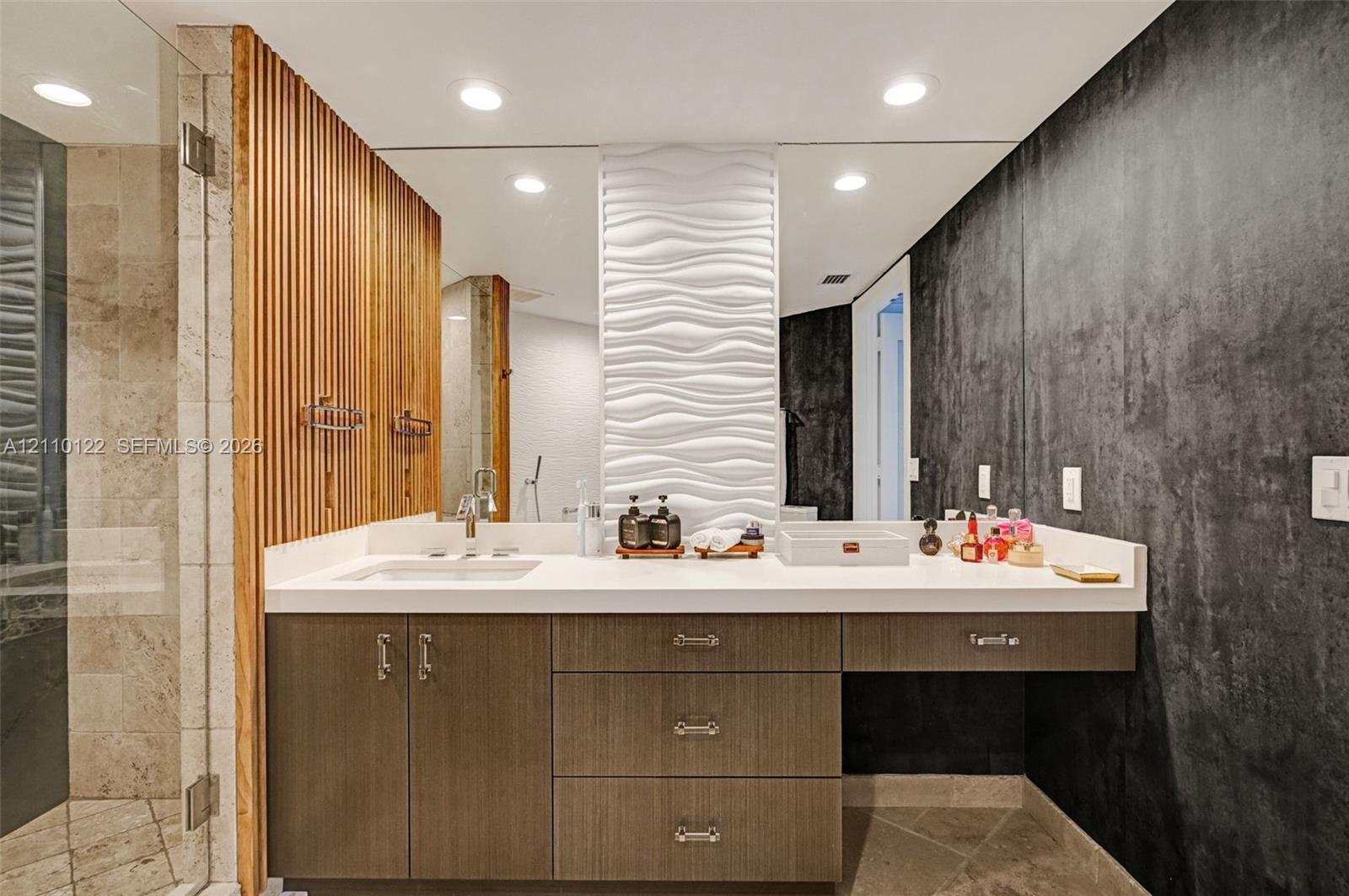 Custom vanity with stone countertop and integrated sink, vertical wood-slat accent wall, 3D wave-pattern wall tile, recessed ceiling lighting, and floor-to-ceiling mirror