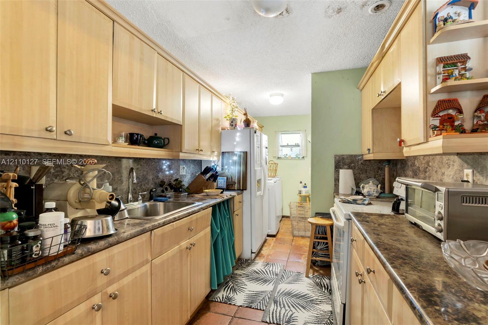 Galley kitchen featuring light wood-finish cabinetry, stone-texture laminate countertops, stainless steel sink with metal faucet, and terra-cotta tile flooring
