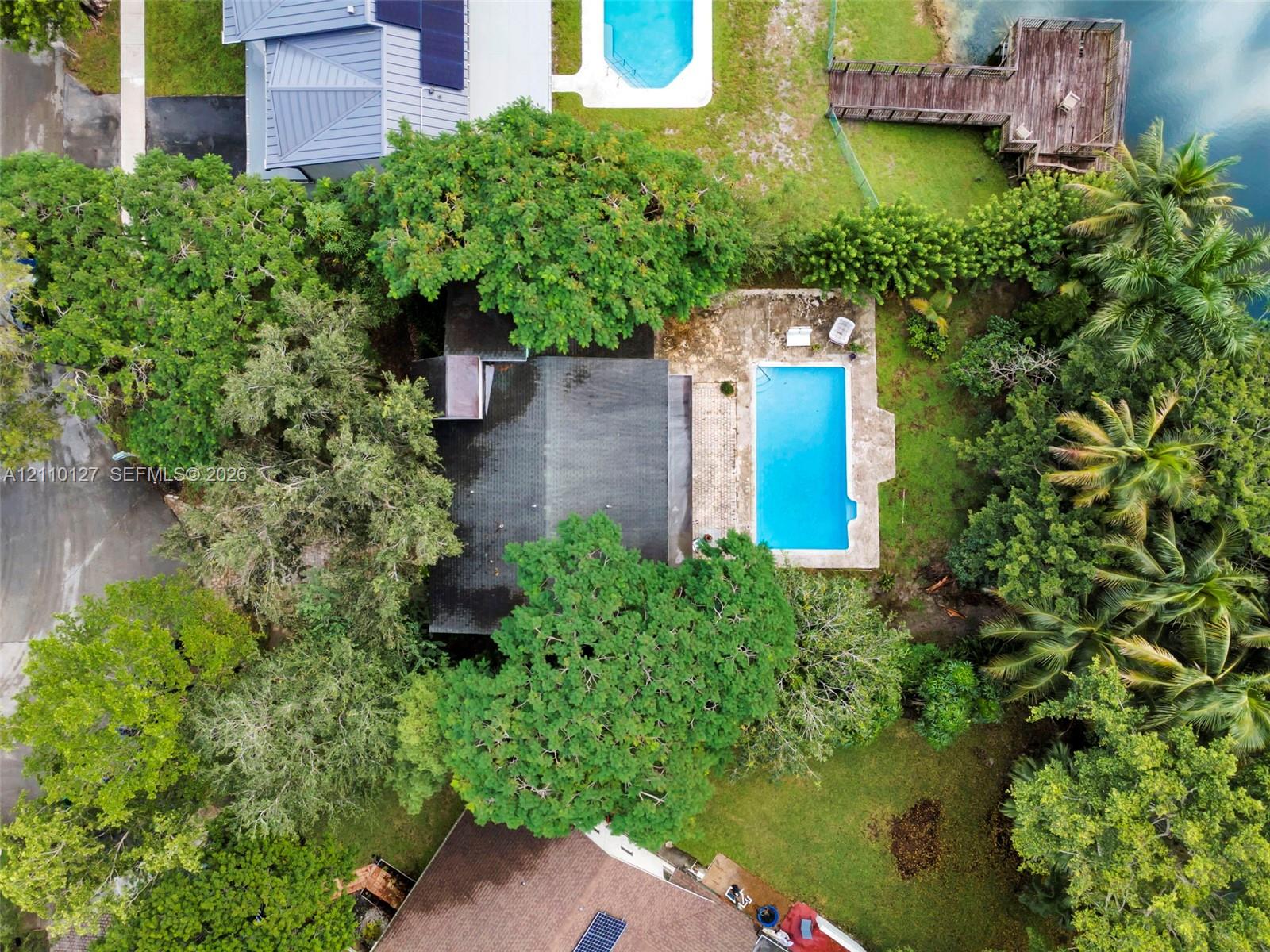Aerial perspective featuring a private swimming pool, stone patio, wooden dock, and mature canopy trees on a waterfront lot Aerial perspective featuring a private swimming pool, stone patio, wooden dock, and mature canopy trees on a waterfront lot