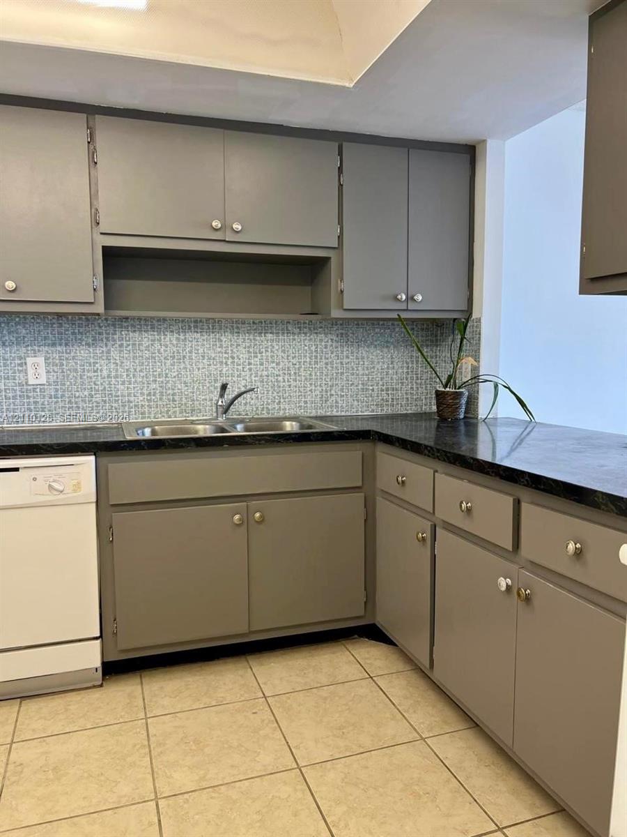 Kitchen featuring gray cabinetry, dark countertops, patterned tile backsplash, dual-basin stainless steel sink, and ceramic tile flooring