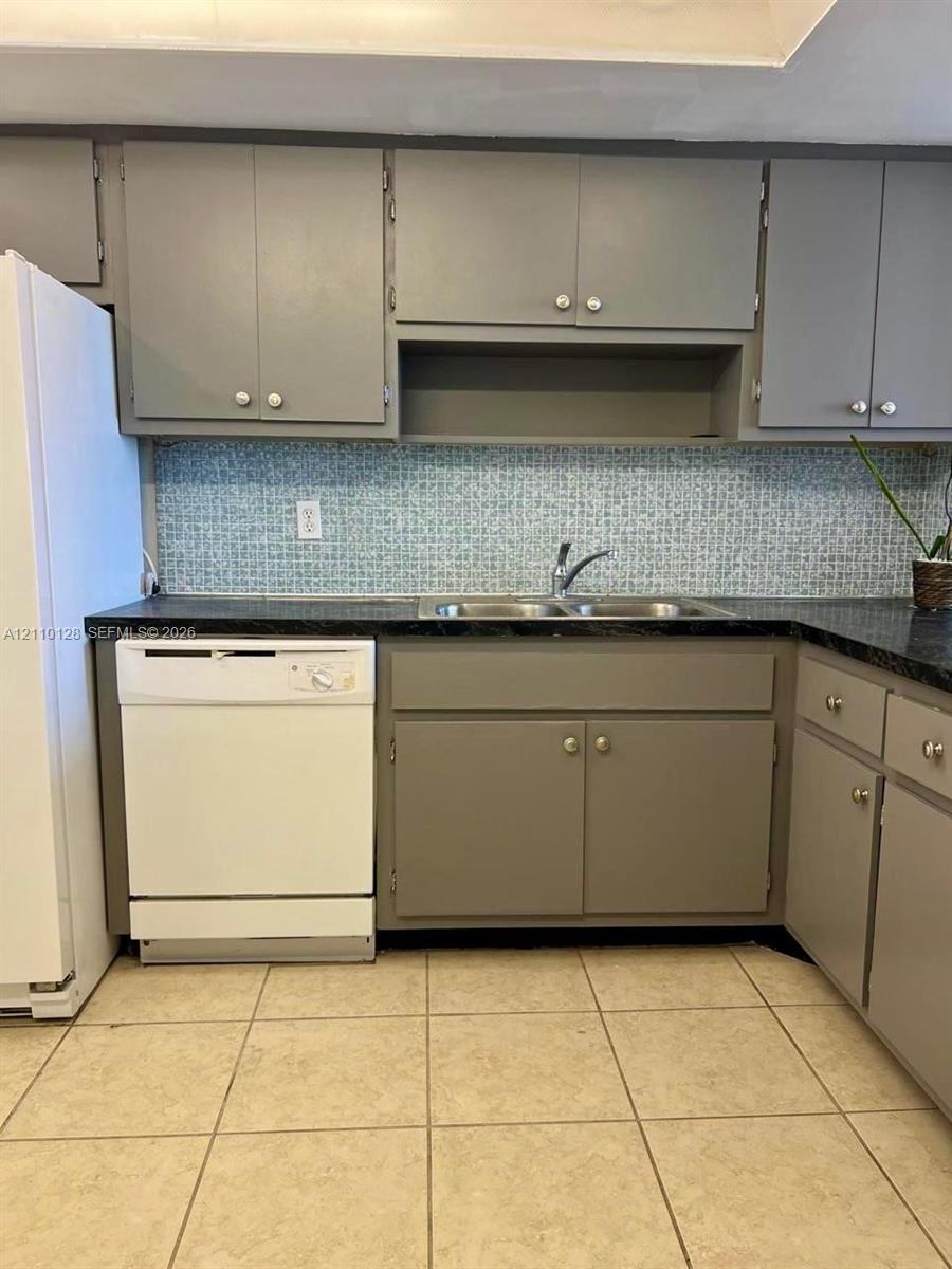 Kitchen featuring gray cabinetry, dark solid-surface countertops, mosaic tile backsplash, double-basin stainless steel sink, and ceramic tile flooring
