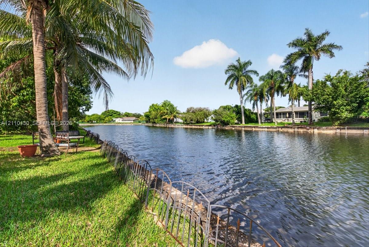 Waterfront property featuring a manicured lawn, metal perimeter fencing, and mature palm trees along the canal bank