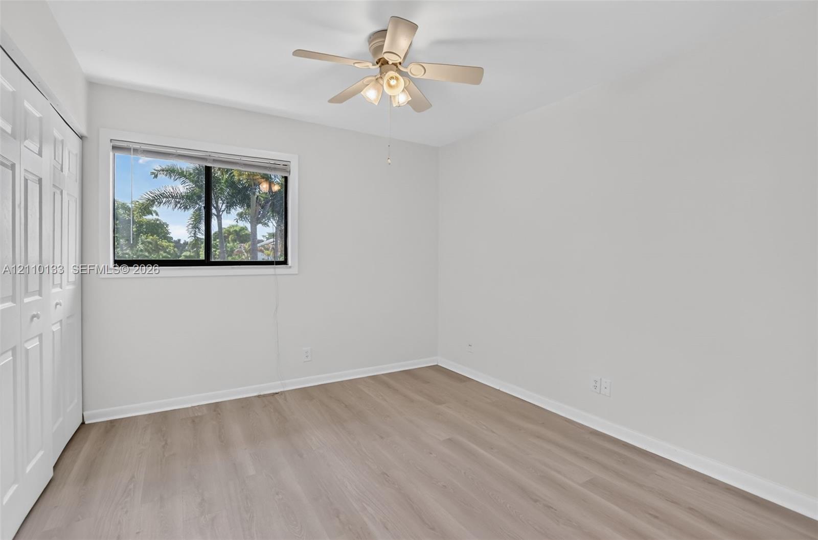 Neutral-toned interior featuring wood-finish flooring, a sliding bi-fold closet, a ceiling fan with multi-light fixture, and a window with exterior greenery