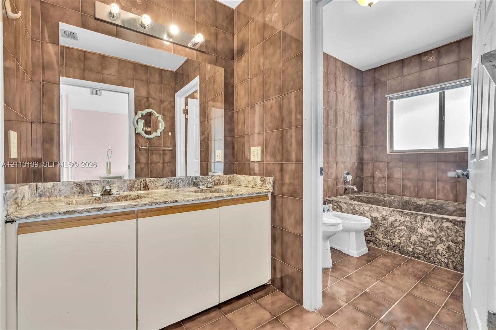 Bathroom featuring a dual-sink vanity with stone-finish countertop, full-wall tiling, a built-in stone-finish tub, and a bidet