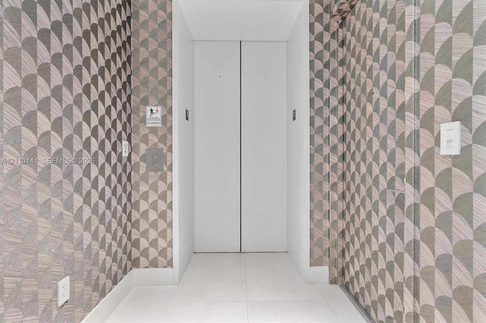Private elevator entry featuring white double doors, geometric patterned wall covering, and neutral tile flooring