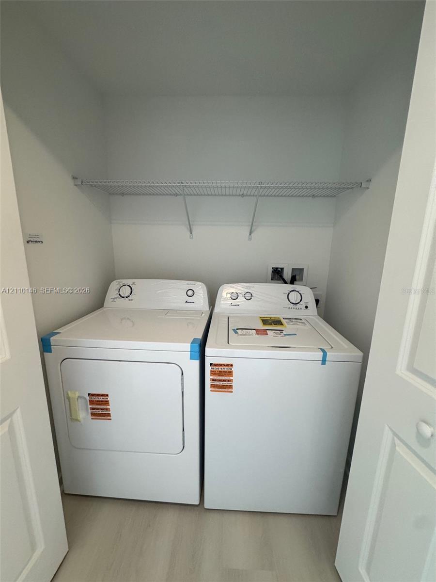 Laundry closet featuring side-by-side white washer and dryer units, a white wire shelf, and light-toned flooring Laundry closet featuring side-by-side white washer and dryer units, a white wire shelf, and light-toned flooring