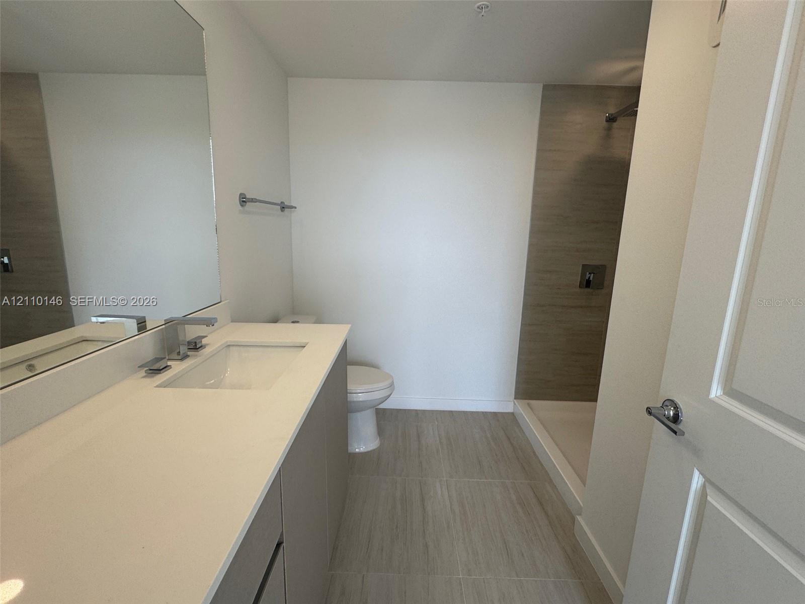 Bathroom featuring a vanity with a white countertop and rectangular sink, large wall-mounted mirror, walk-in shower with neutral tile surround, and tile flooring