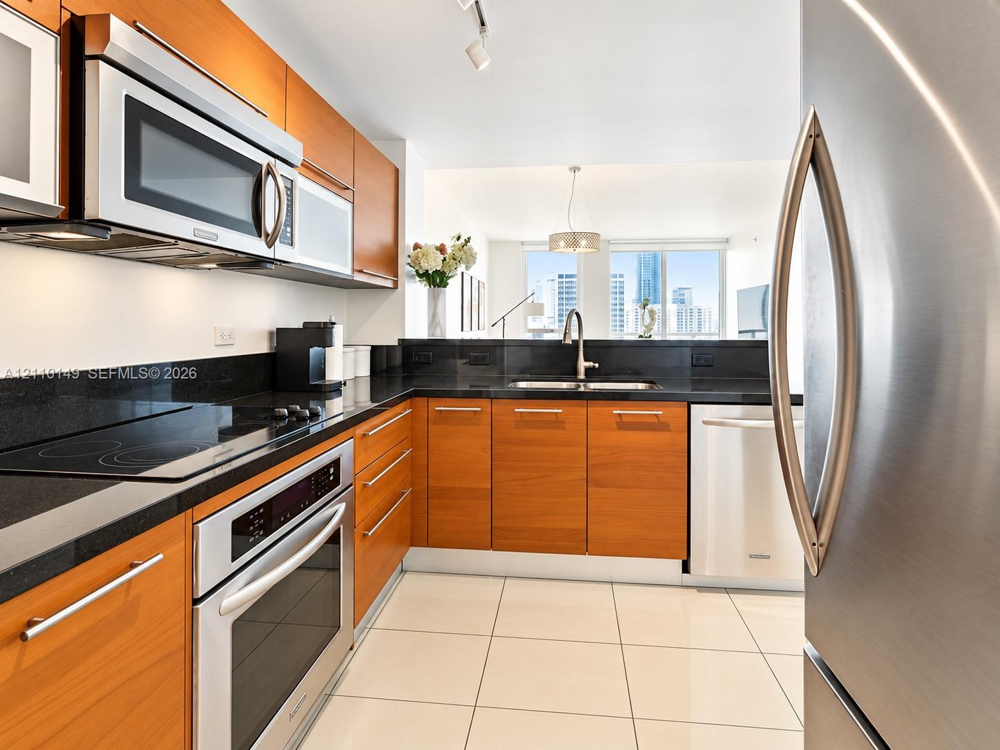 Kitchen featuring wood-finish cabinetry, dark stone countertops, stainless steel appliances, and large-format tile flooring Kitchen featuring wood-finish cabinetry, dark stone countertops, stainless steel appliances, and large-format tile flooring