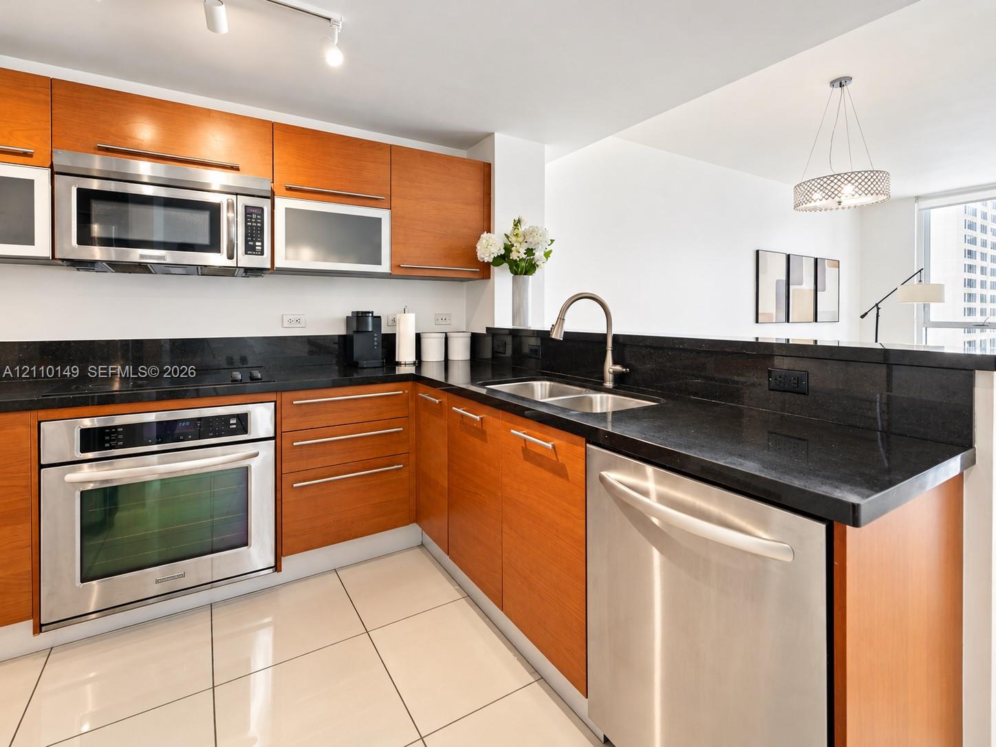 Kitchen featuring wood-finish cabinetry, dark stone countertops, stainless steel appliances, dual-basin sink, and tile flooring Kitchen featuring wood-finish cabinetry, dark stone countertops, stainless steel appliances, dual-basin sink, and tile flooring