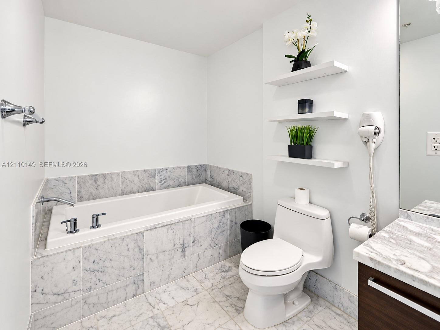 Bathroom featuring a stone-surround soaking tub, stone tile flooring, a vanity with a stone countertop, and three integrated wall-mounted shelves Bathroom featuring a stone-surround soaking tub, stone tile flooring, a vanity with a stone countertop, and three integrated wall-mounted shelves