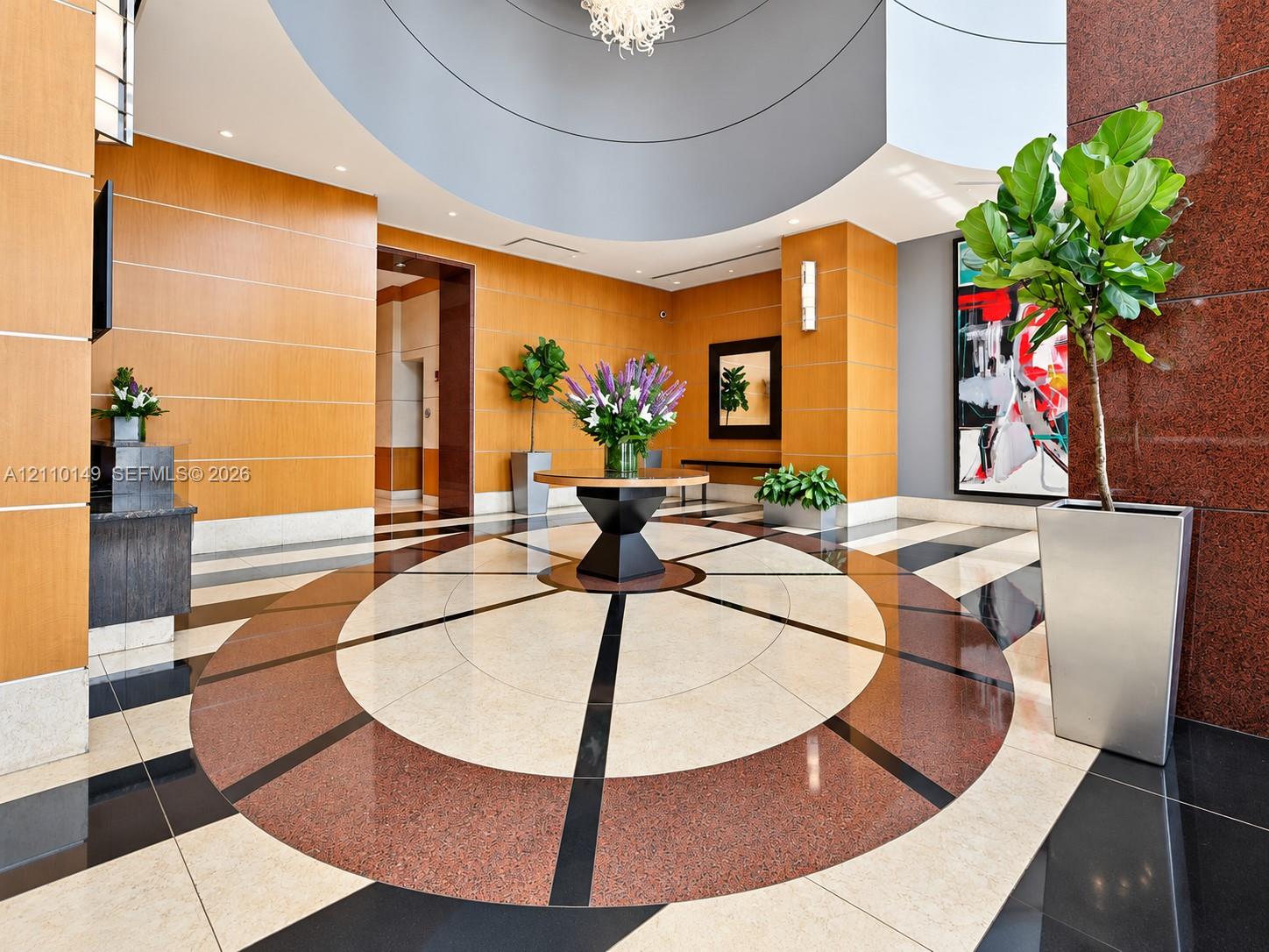 Grand foyer featuring geometric stone flooring, warm wood-paneled walls, a high circular ceiling, polished red stone accents, and a contemporary multi-arm chandelier