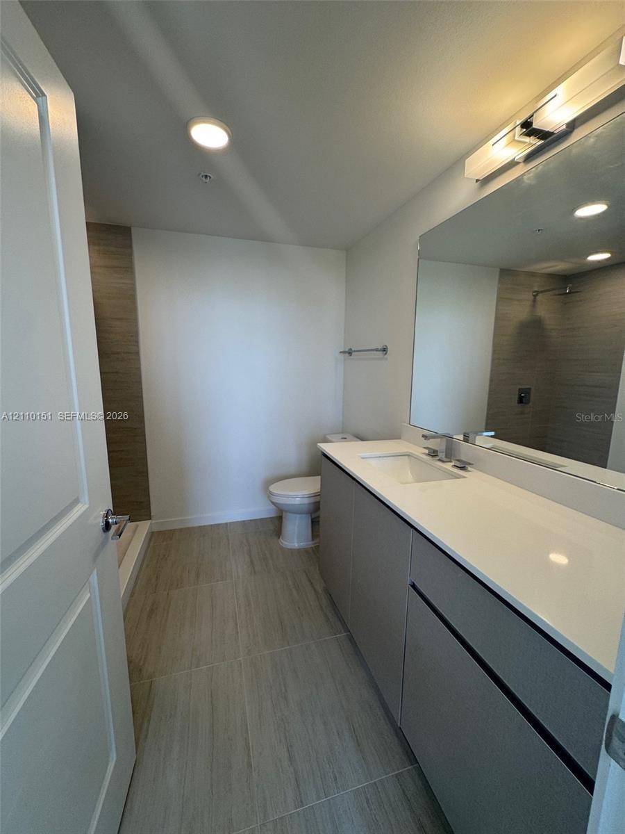 Bathroom featuring a floating vanity with a light-toned countertop, large wall-mounted mirror, wood-finish floor tiling, and a walk-in shower with vertical tile surround