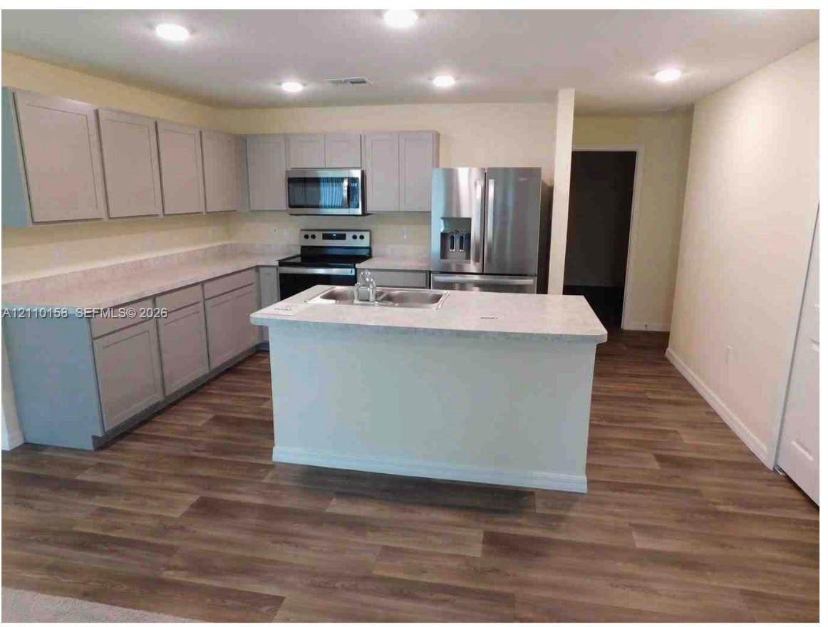 Kitchen featuring light-toned cabinetry, stone-finish countertops, stainless steel appliances, a center island with a dual-basin sink, and wood-finish flooring Kitchen featuring light-toned cabinetry, stone-finish countertops, stainless steel appliances, a center island with a dual-basin sink, and wood-finish flooring
