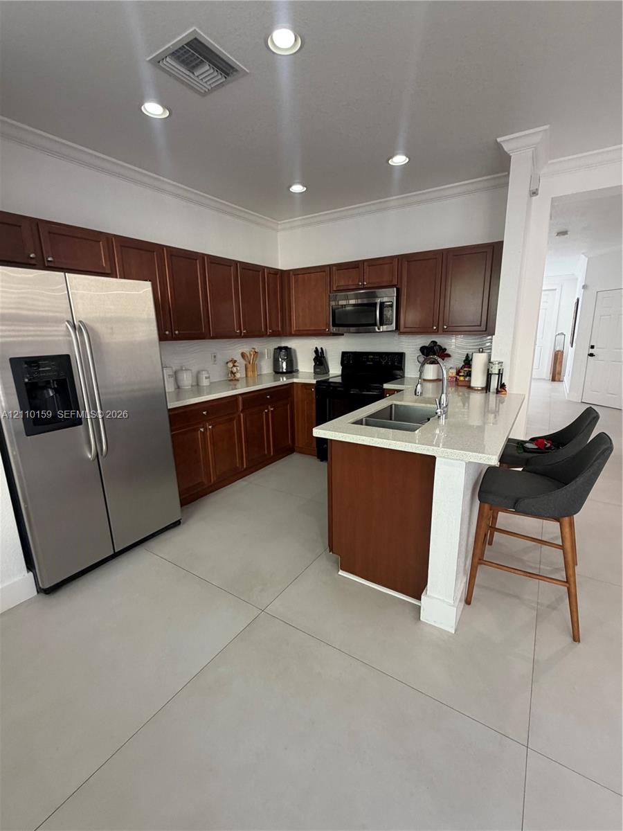 Kitchen featuring wood cabinetry, light-toned stone countertops, a center island with a dual-basin stainless steel sink, large-format tile flooring, and recessed ceiling lighting