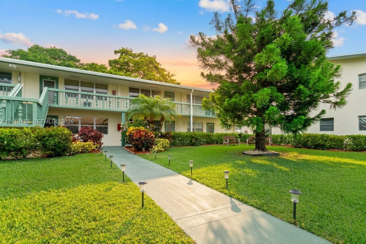 Two-story exterior featuring painted stucco facade, second-level balcony with railings, concrete pedestrian walkway, manicured lawn, and mature landscaping Two-story exterior featuring painted stucco facade, second-level balcony with railings, concrete pedestrian walkway, manicured lawn, and mature landscaping