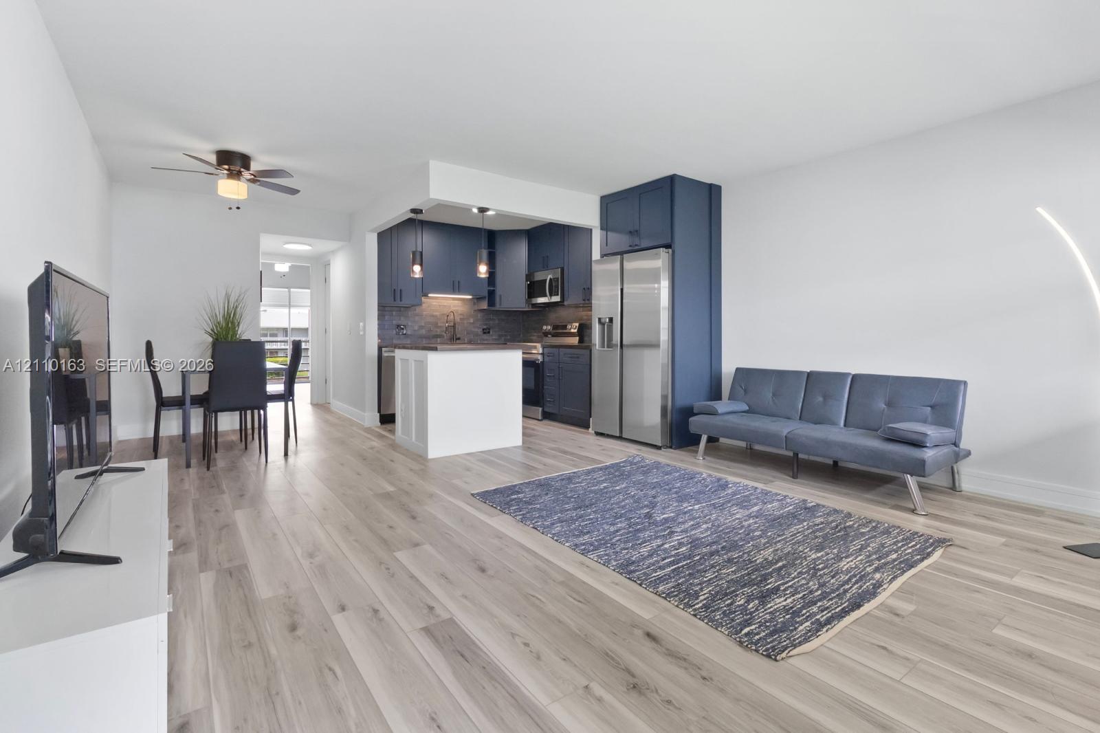 Open-concept space featuring wood-finish flooring, navy-blue kitchen cabinetry, stainless steel appliances, a white center island, and dual pendant lighting