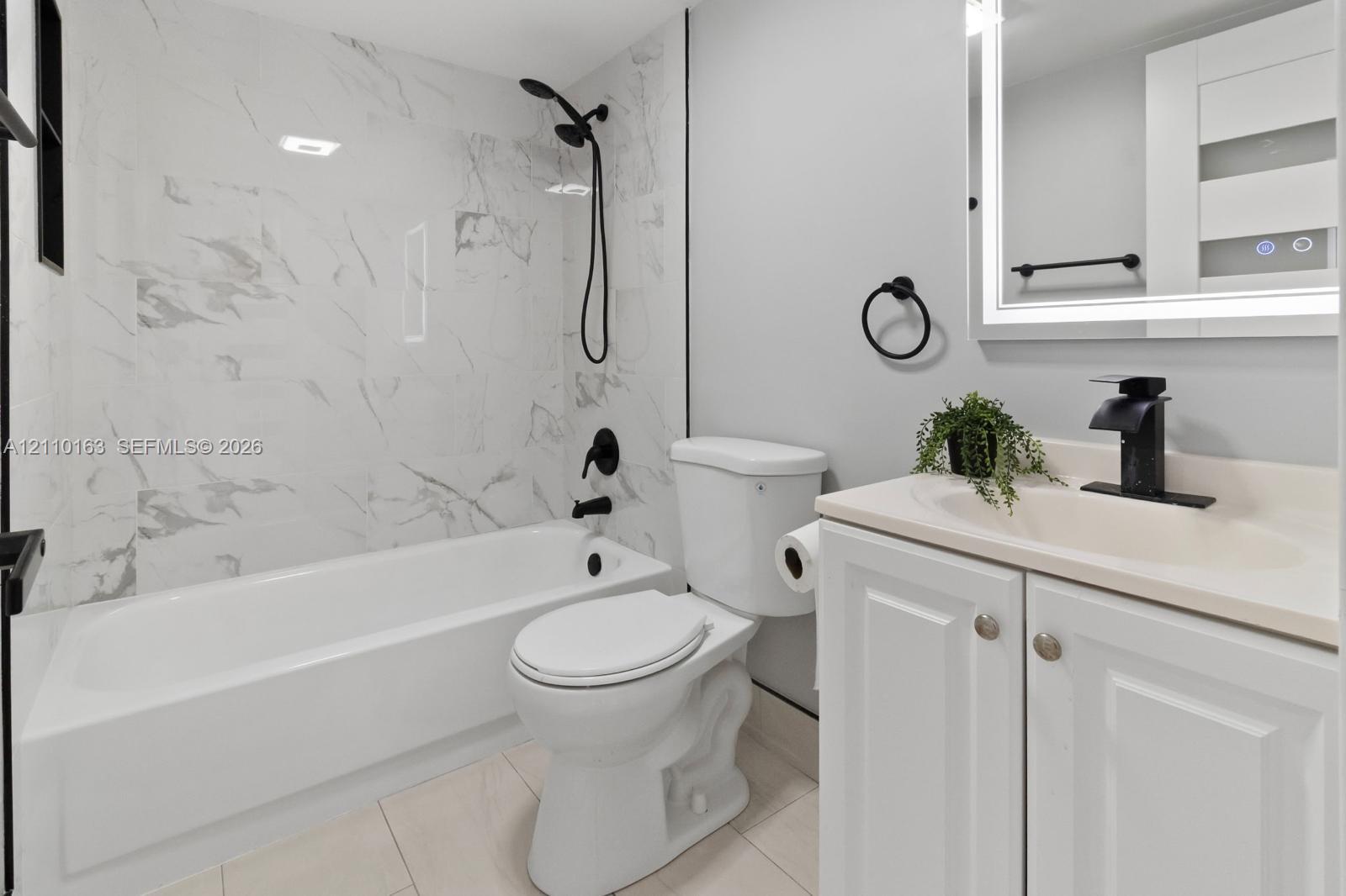 Bathroom featuring a soaking tub with full-height stone-look tile surround, vanity with an integrated sink, matte black faucet hardware, and a backlit mirror
