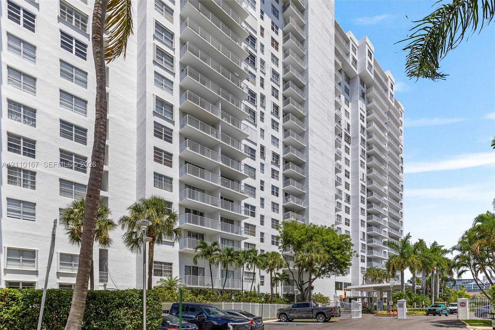 High-rise residential building featuring white stucco facade, stacked private balconies, covered porte-cochre entry, and landscaped tropical grounds with mature palm trees