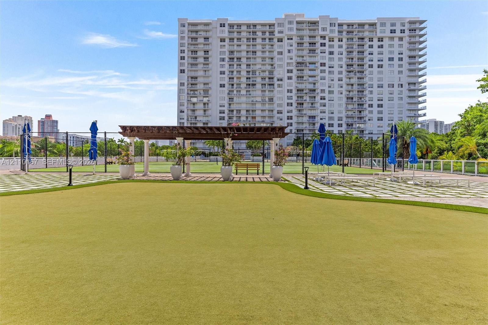 Synthetic turf putting green featuring a wood-beam pergola, stone-tile hardscaping, and perimeter fencing