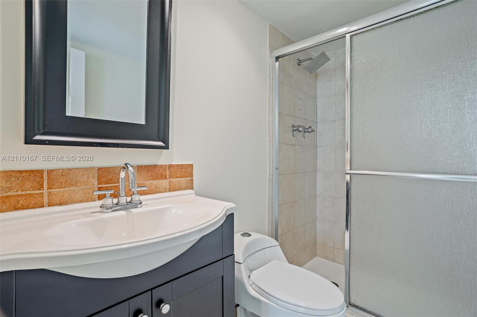 Dark finish vanity with integrated sink, chrome faucet, framed mirror, tiled backsplash, and glass-enclosed shower stall