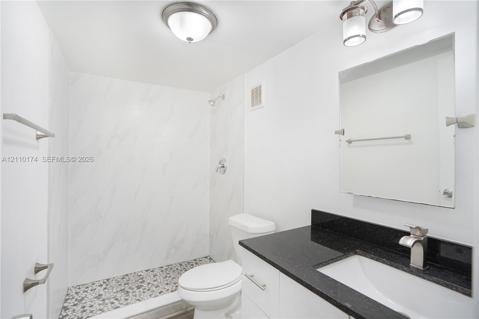Full-height shower featuring large-format stone-pattern wall tiles and a decorative pebble-style basin floor, vanity with a solid-surface countertop and undermount sink, and wall-mounted vanity mirror with integrated lighting