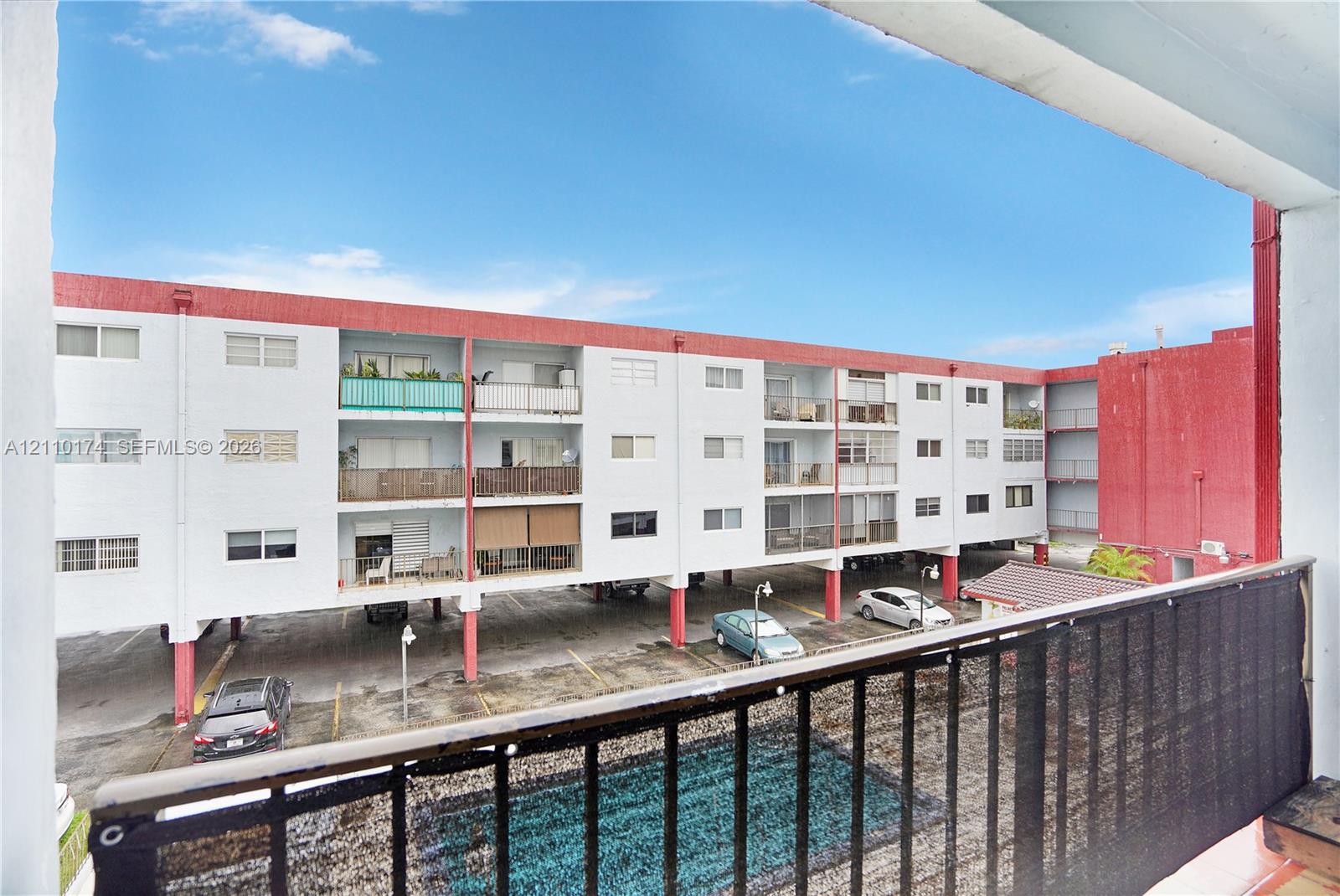 Exterior balcony featuring black metal railings and a privacy screen, overlooking a ground-level swimming pool and covered parking area