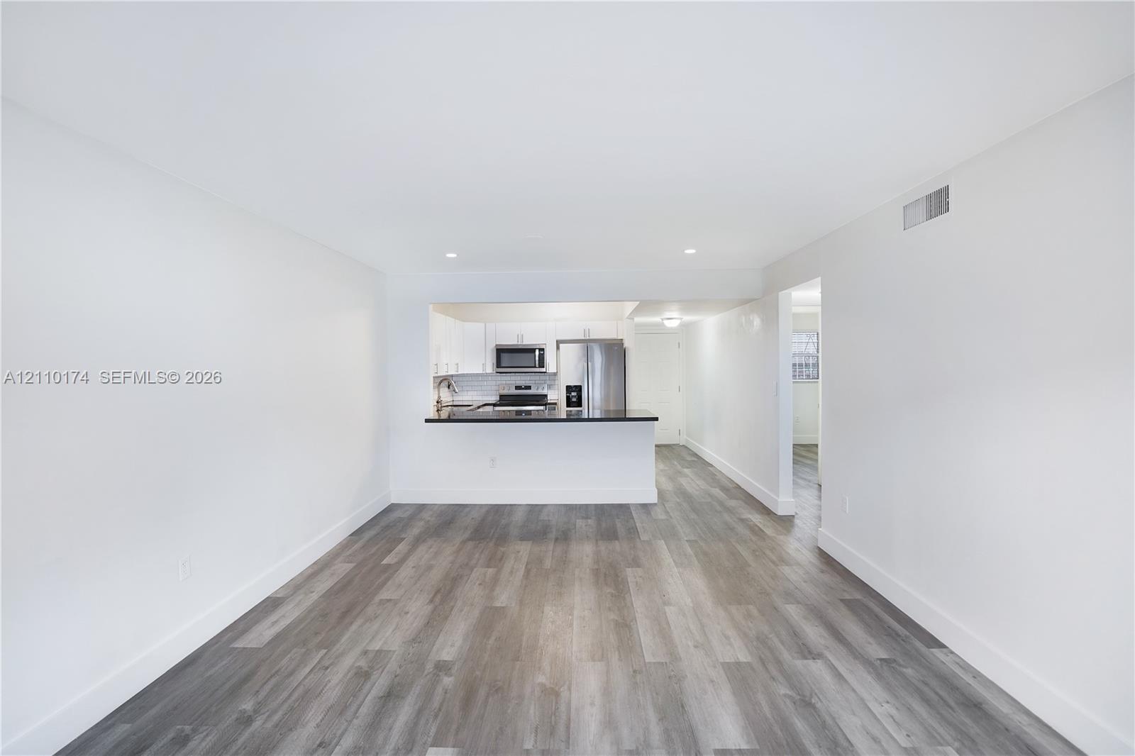 Open-concept interior featuring wood-finish flooring, white cabinetry, dark countertops, and stainless steel kitchen appliances Open-concept interior featuring wood-finish flooring, white cabinetry, dark countertops, and stainless steel kitchen appliances