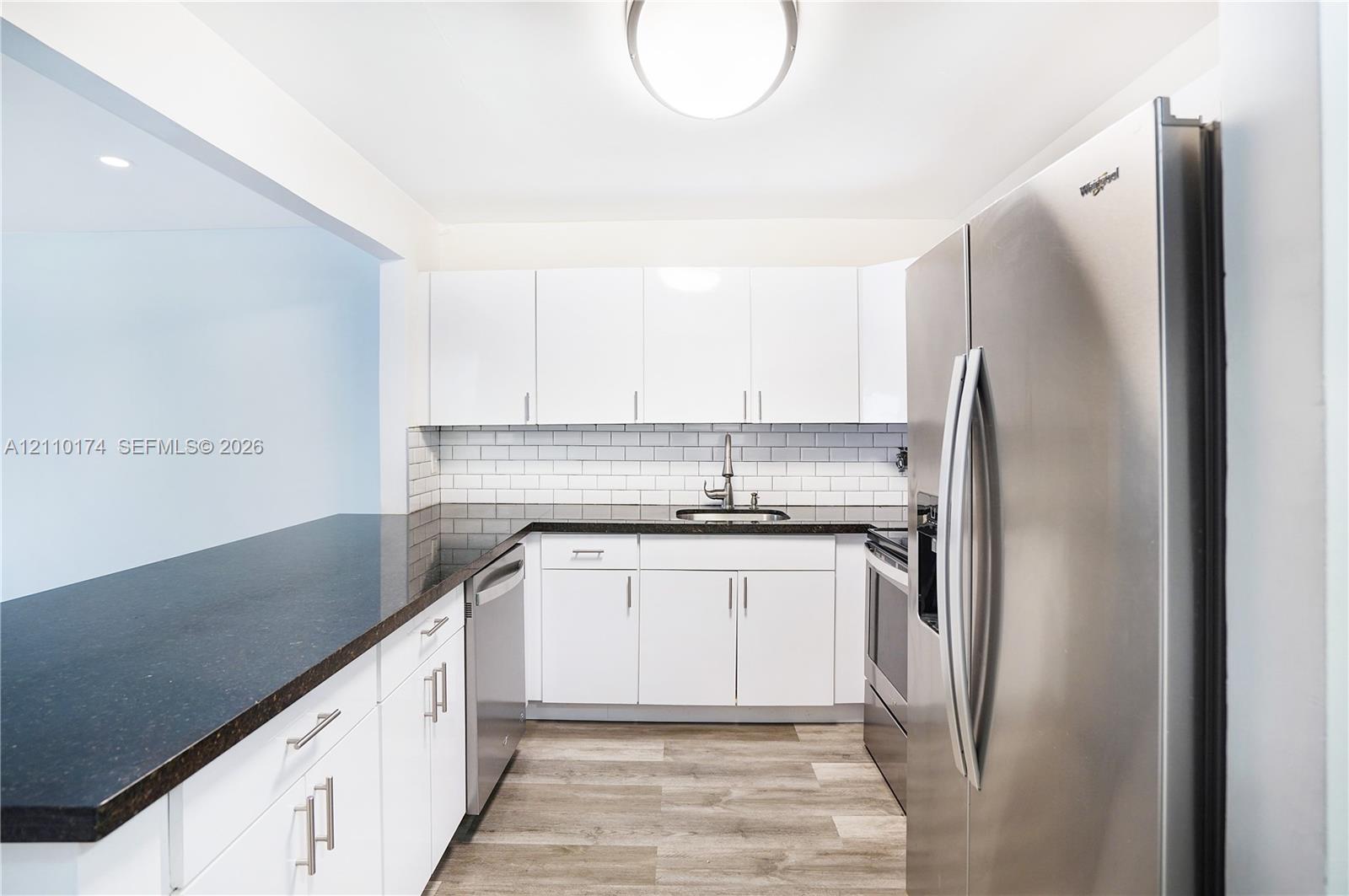 Kitchen featuring white flat-panel cabinetry, dark stone-finish countertops, stainless steel appliances, a white subway tile backsplash, and wood-finish flooring Kitchen featuring white flat-panel cabinetry, dark stone-finish countertops, stainless steel appliances, a white subway tile backsplash, and wood-finish flooring