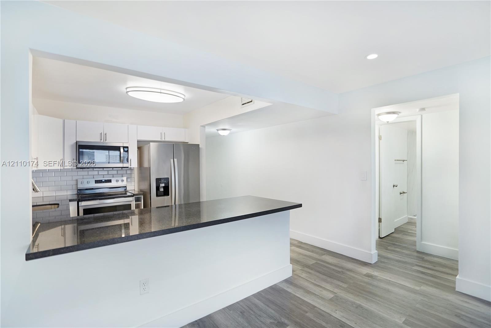 Kitchen featuring a dark stone countertop peninsula, white shaker-style cabinetry, stainless steel appliances, grey subway tile backsplash, and wood-finish flooring Kitchen featuring a dark stone countertop peninsula, white shaker-style cabinetry, stainless steel appliances, grey subway tile backsplash, and wood-finish flooring