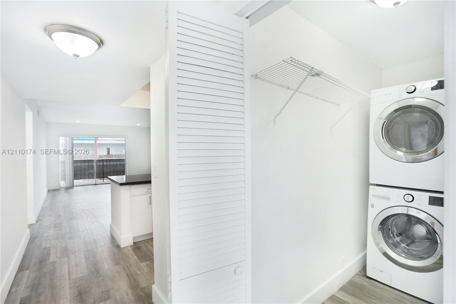 Laundry area featuring a stacked washer and dryer unit, integrated wire shelving, wood-finish flooring, and a louvered bi-fold door Laundry area featuring a stacked washer and dryer unit, integrated wire shelving, wood-finish flooring, and a louvered bi-fold door