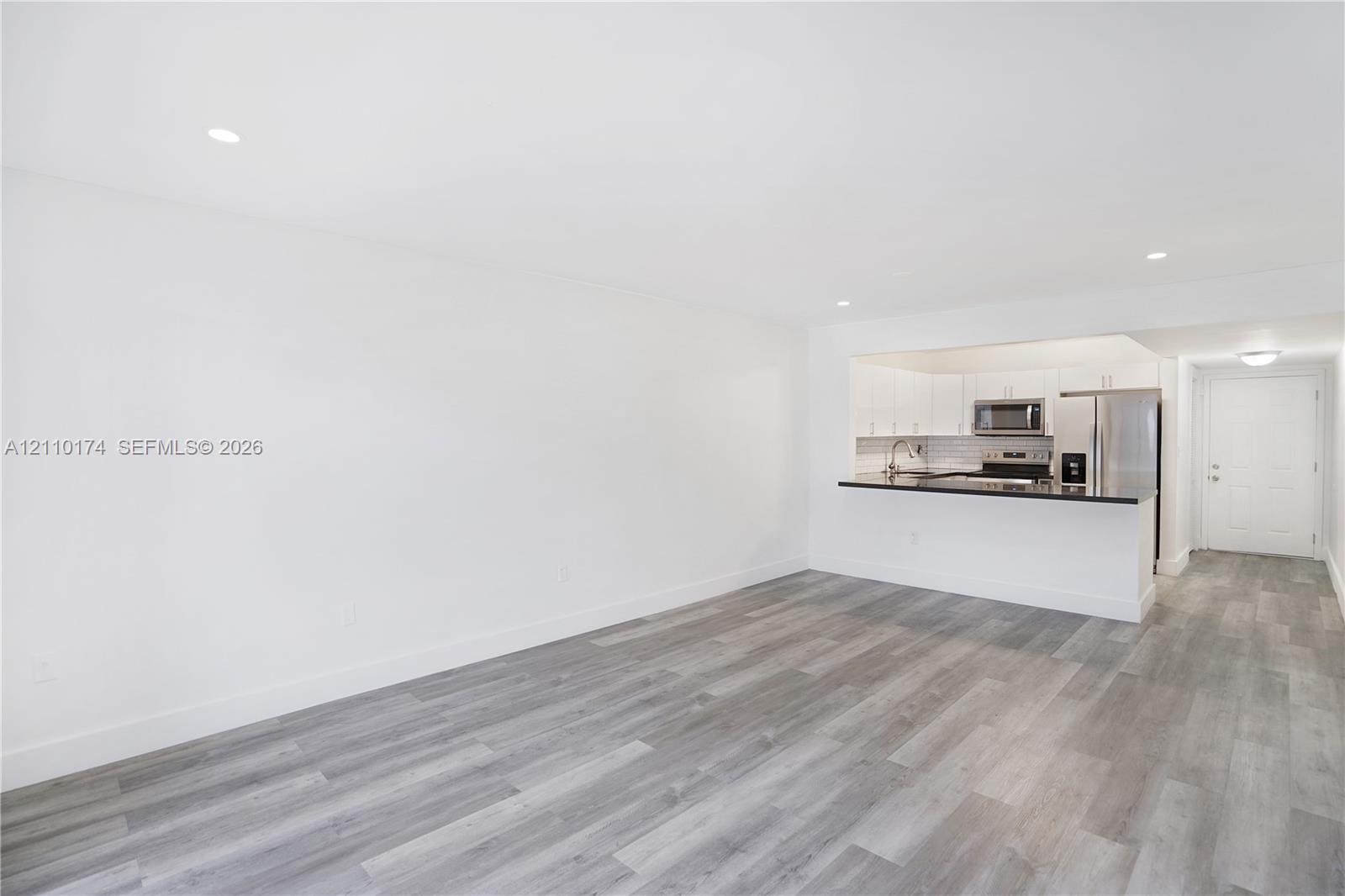 Open-concept interior featuring wood-finish flooring, recessed lighting, and a kitchen with white cabinetry, stainless steel appliances, and a dark-toned countertop breakfast bar Open-concept interior featuring wood-finish flooring, recessed lighting, and a kitchen with white cabinetry, stainless steel appliances, and a dark-toned countertop breakfast bar