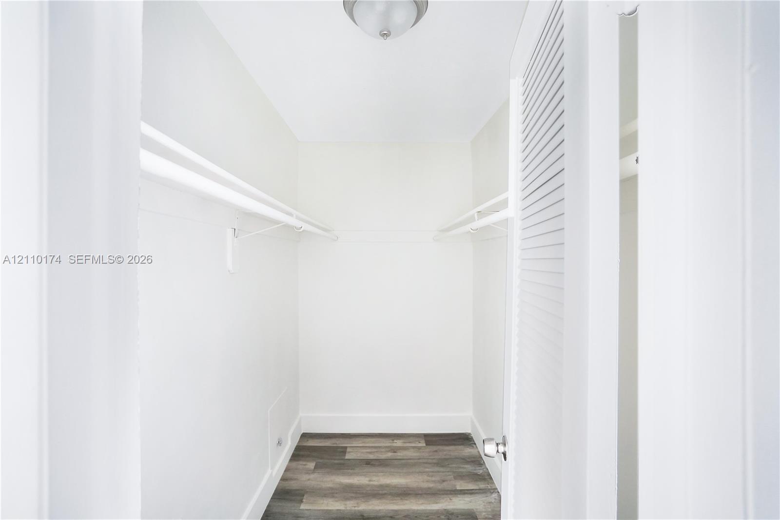 Storage area featuring wood-finish flooring, dual-level wall-mounted shelving, and a louvered door