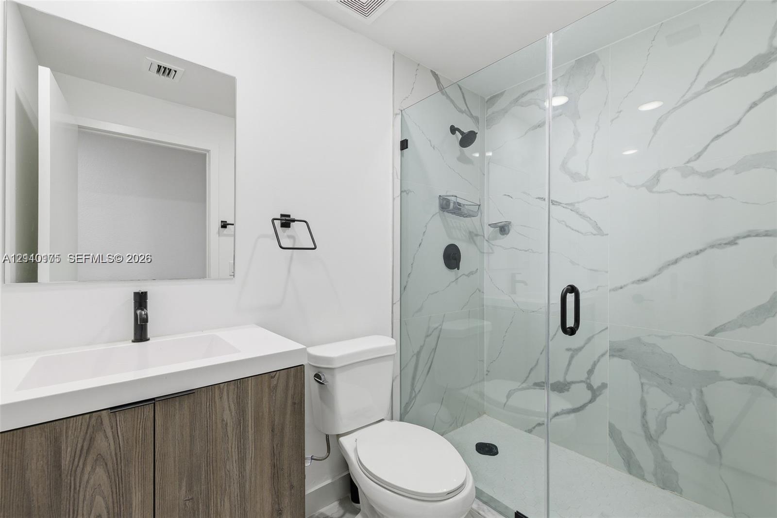 Wood-finish cabinetry with a white countertop, rectangular vessel sink, wall-mounted matte black faucet, and walk-in shower with floor-to-ceiling stone-patterned tile and a frameless glass door