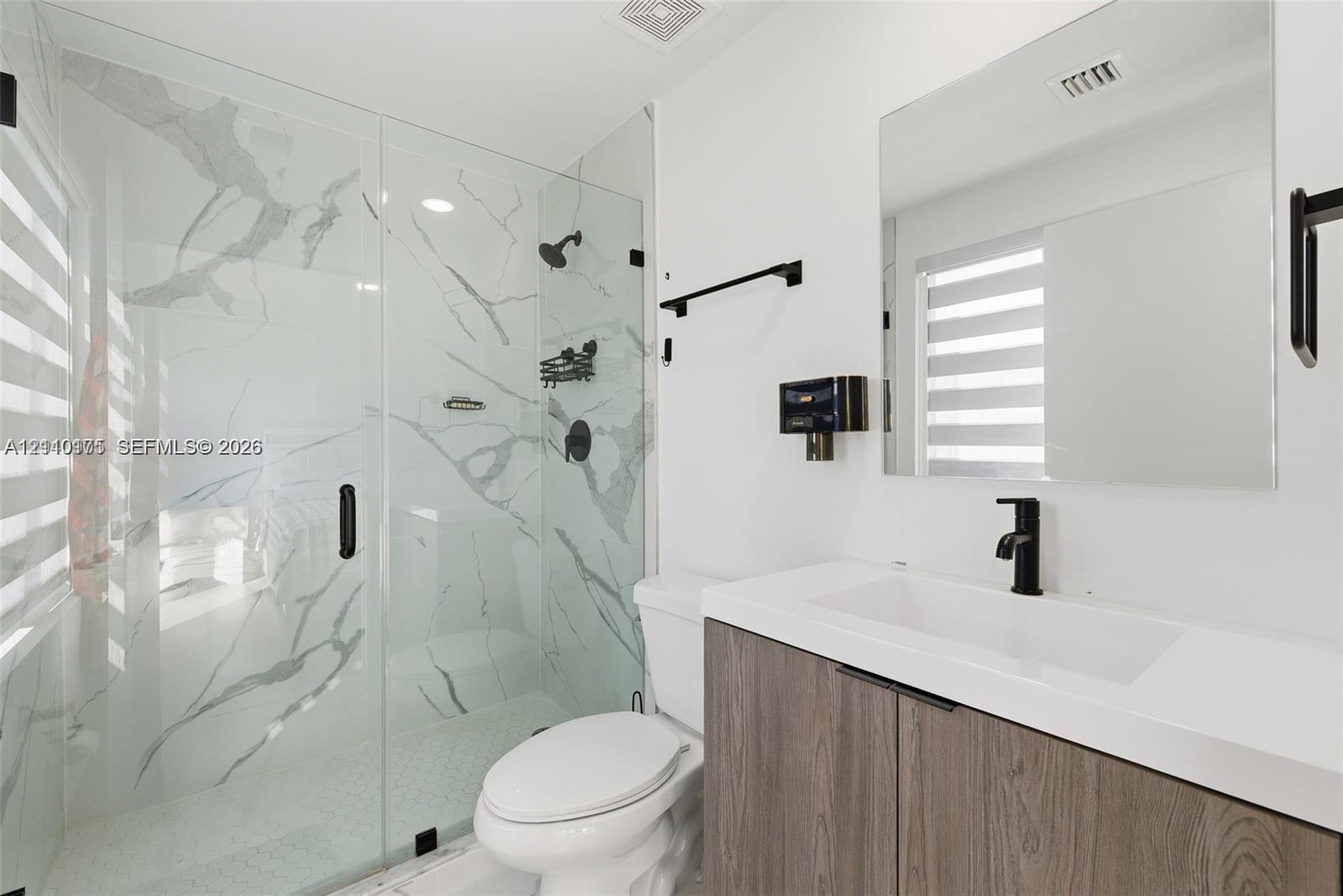 Walk-in shower with stone-finish tile surround and glass enclosure, wood-finish floating vanity with white countertop, matte black fixtures, and large frameless mirror