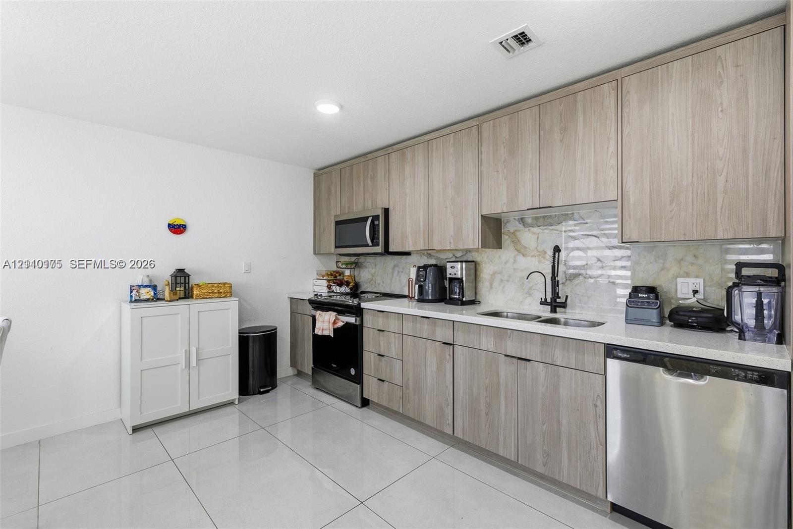 Kitchen featuring flat-panel wood-finish cabinetry, light-toned stone-surface countertops, stone tile backsplash, stainless steel appliances, and large-format porcelain tile flooring