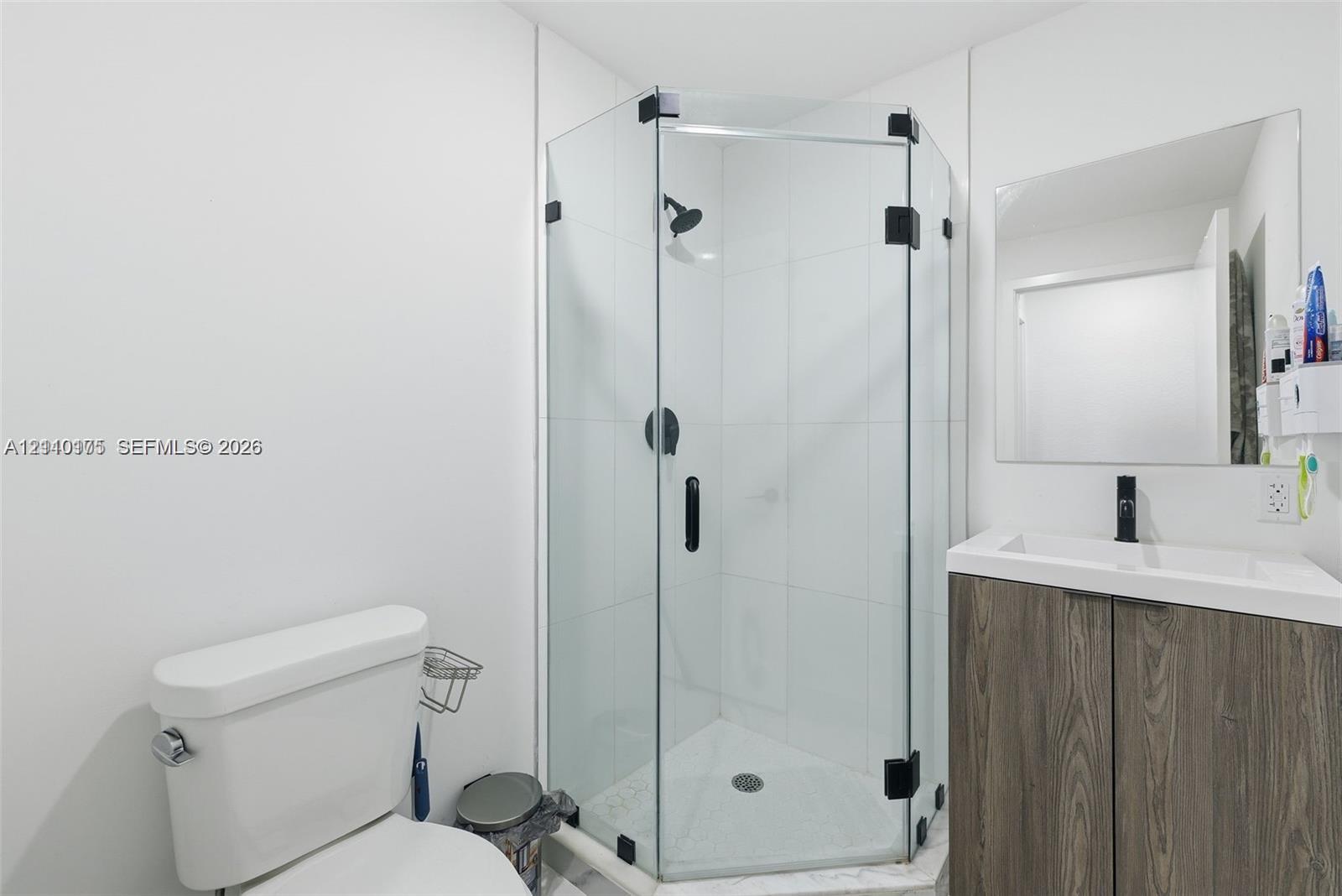 Corner shower enclosure with glass panels, wood-finish vanity with integrated sink, matte black hardware, and wall-mounted mirror