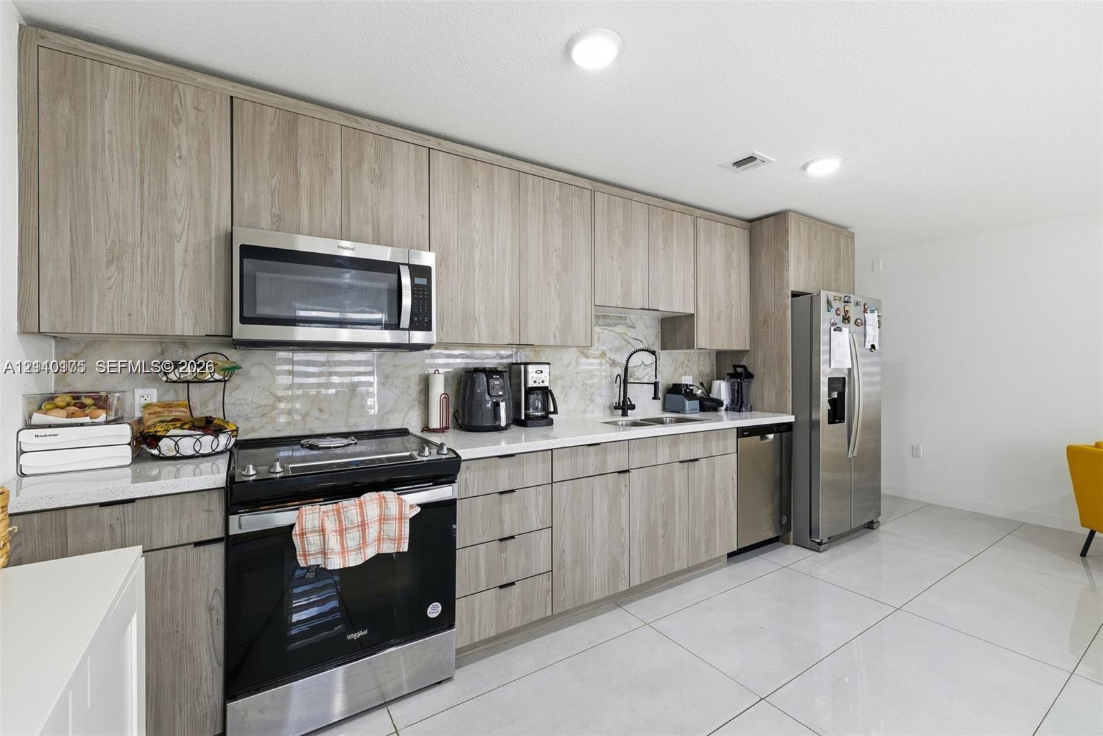 Kitchen featuring wood-finish cabinetry, stainless steel appliances, stone-surface backsplash, and large-format tile flooring