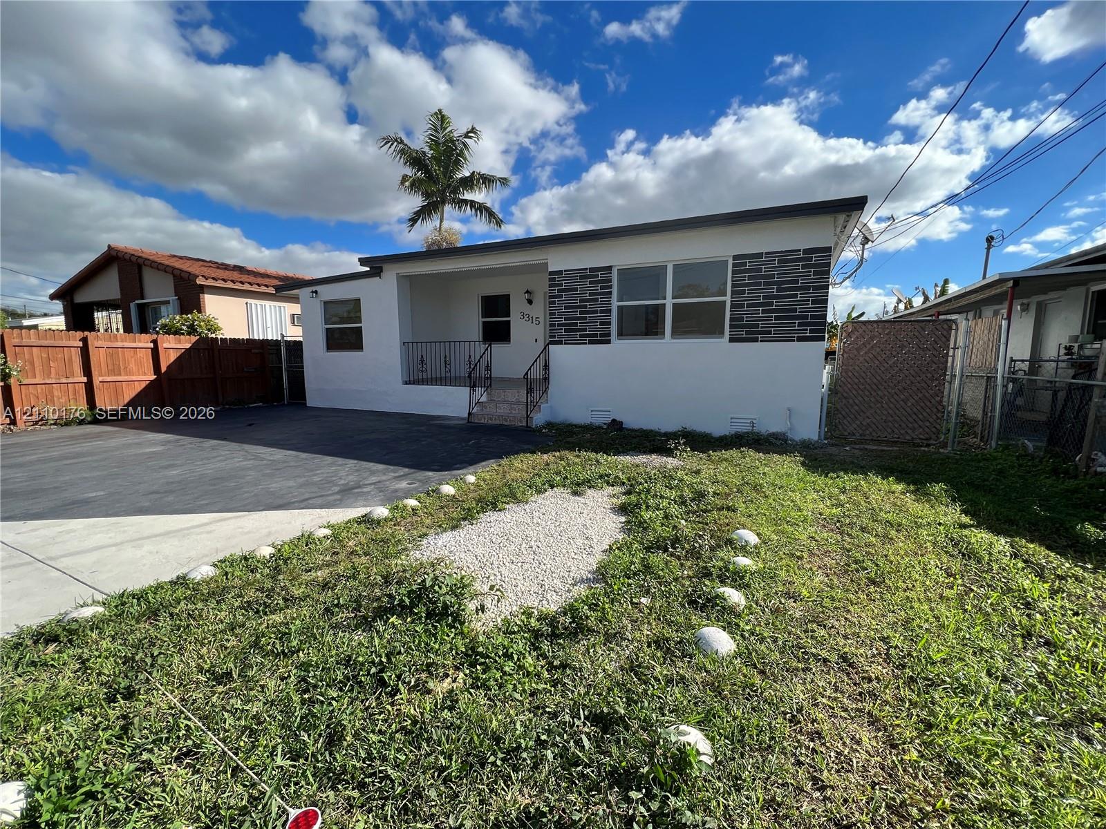 Single-story exterior featuring white stucco finish, decorative brick masonry detailing, covered front entryway, metal railings, and paved driveway