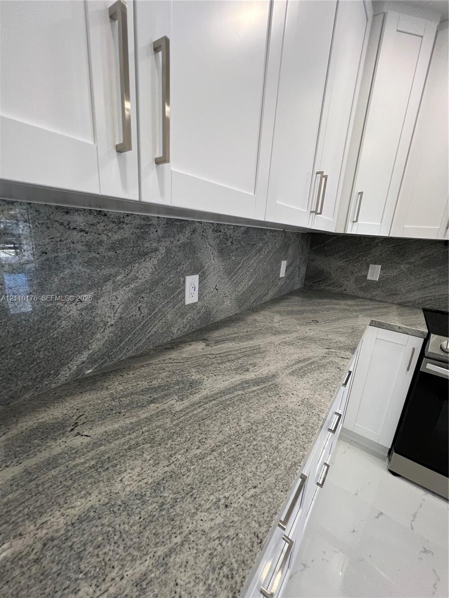 Kitchen featuring white shaker-style cabinetry, stone countertops, matching full-height stone backsplash, and stainless steel hardware