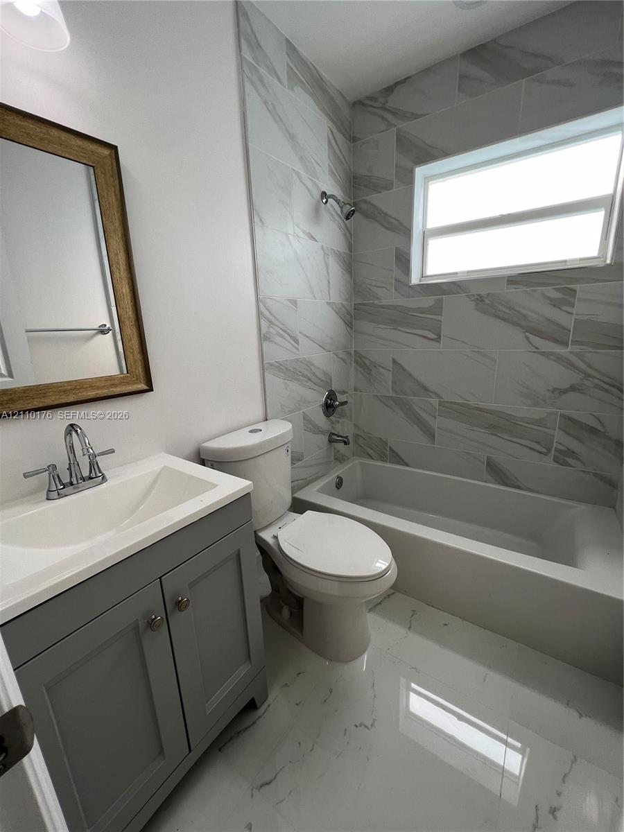 Bathroom featuring gray shaker-style vanity cabinetry, white integrated sink countertop, large-format tile flooring, and tub-shower combination with full-height wall tiling