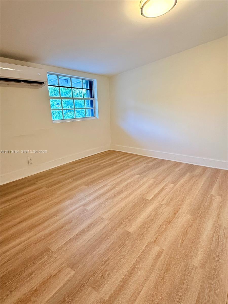 Room featuring wood-finish flooring, a wall-mounted ductless air conditioning unit, a multi-pane window, and a flush-mount ceiling light fixture Room featuring wood-finish flooring, a wall-mounted ductless air conditioning unit, a multi-pane window, and a flush-mount ceiling light fixture