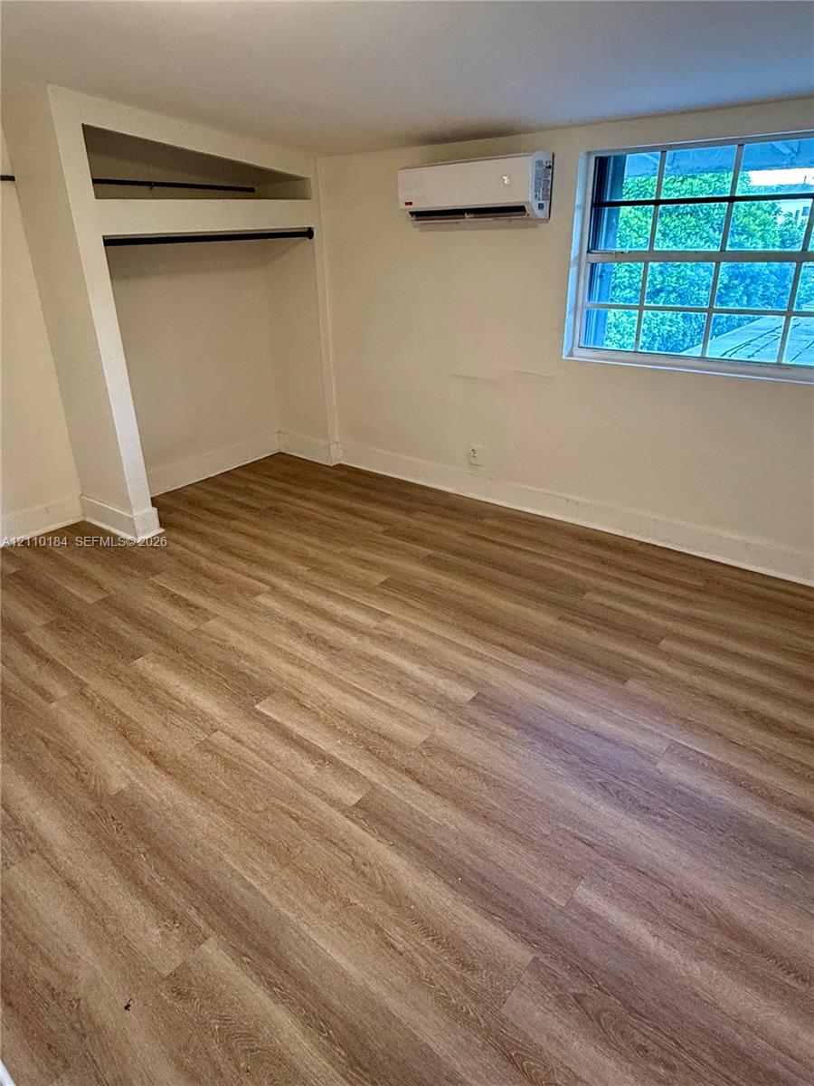 Interior space featuring wood-finish flooring, a wall-mounted ductless mini-split system, a multi-pane window, and a built-in closet with hanging rods Interior space featuring wood-finish flooring, a wall-mounted ductless mini-split system, a multi-pane window, and a built-in closet with hanging rods