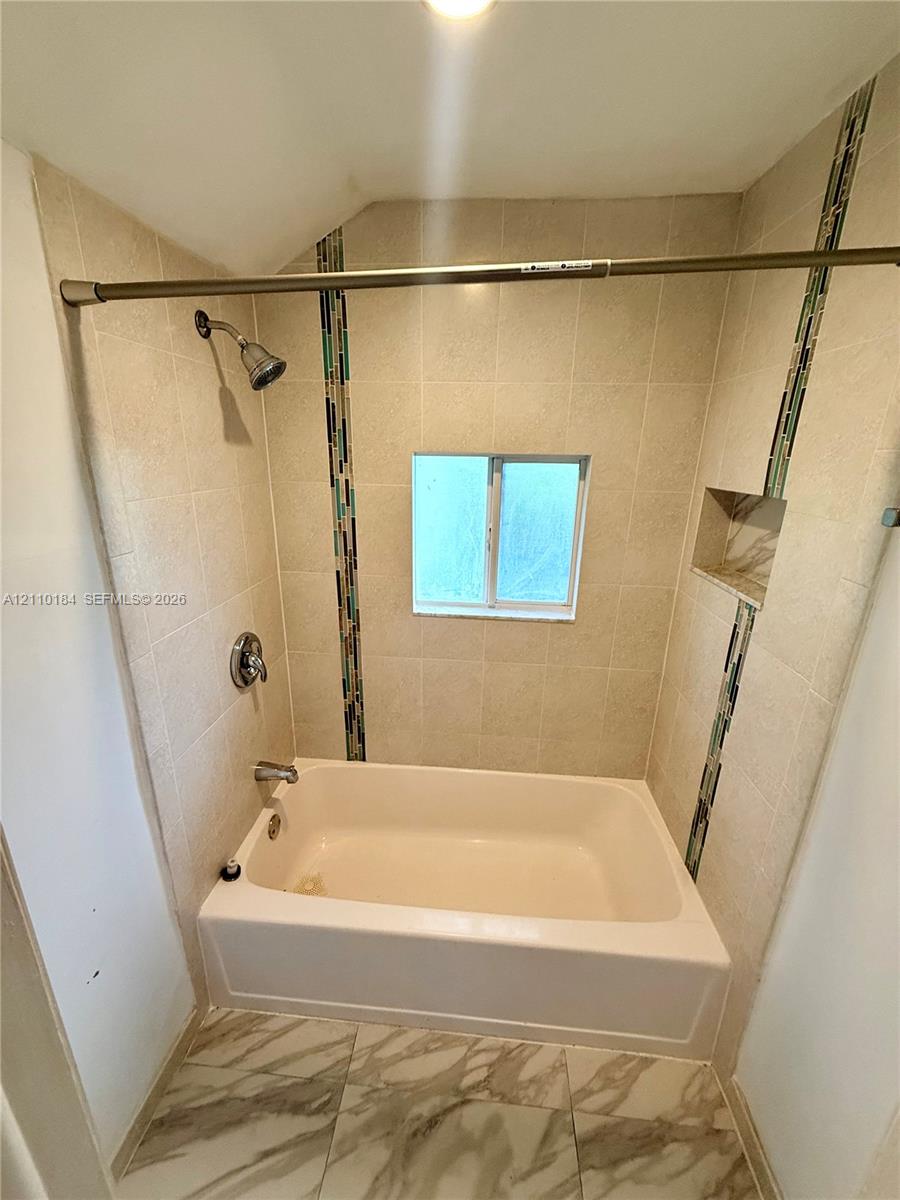Bathtub and shower combination featuring neutral tile surround with decorative glass mosaic vertical accents, built-in recessed storage niche, and stone-finish floor tile Bathtub and shower combination featuring neutral tile surround with decorative glass mosaic vertical accents, built-in recessed storage niche, and stone-finish floor tile