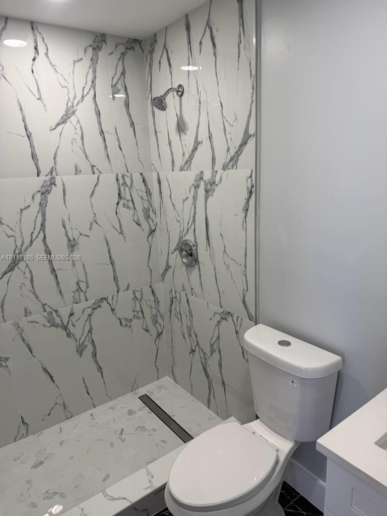 Shower enclosure featuring large-format veined tile surround, chrome shower head, integrated linear drain, and a white finish toilet