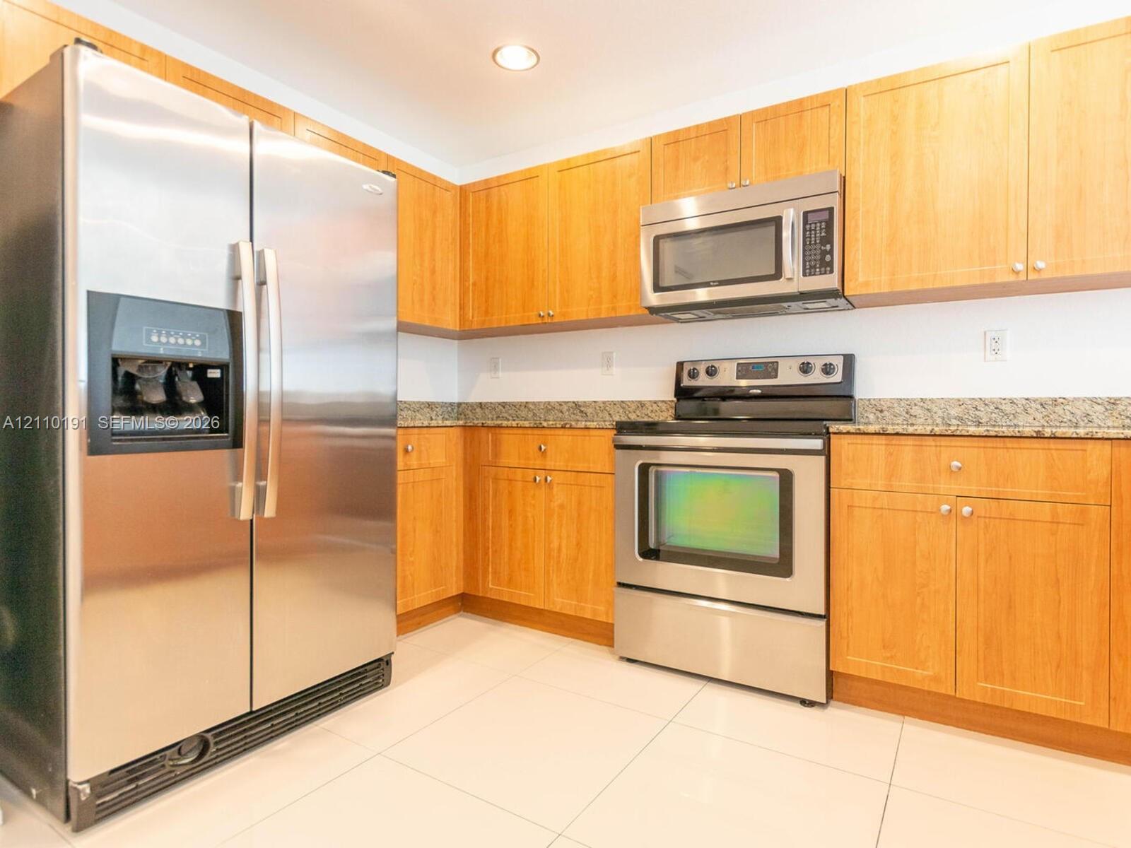 Kitchen featuring natural wood-finish cabinetry, speckled stone countertops, and stainless steel appliances including a double-door refrigerator, over-the-range microwave, and slide-in range with large-format floor tile