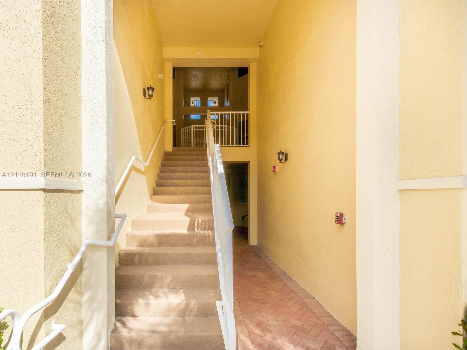 Exterior staircase featuring concrete steps, white metal railings, herringbone-patterned paved walkway, and wall-mounted sconces