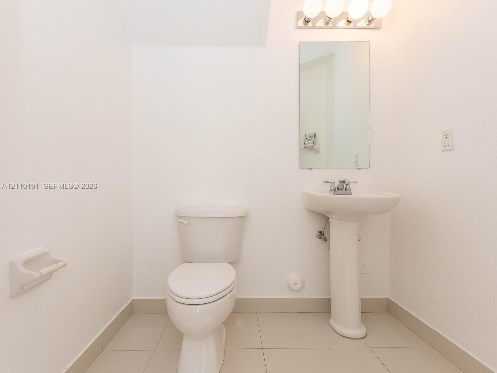 Pedestal sink with chrome faucet, wall-mounted rectangular mirror, four-bulb vanity lighting, and ceramic tile flooring