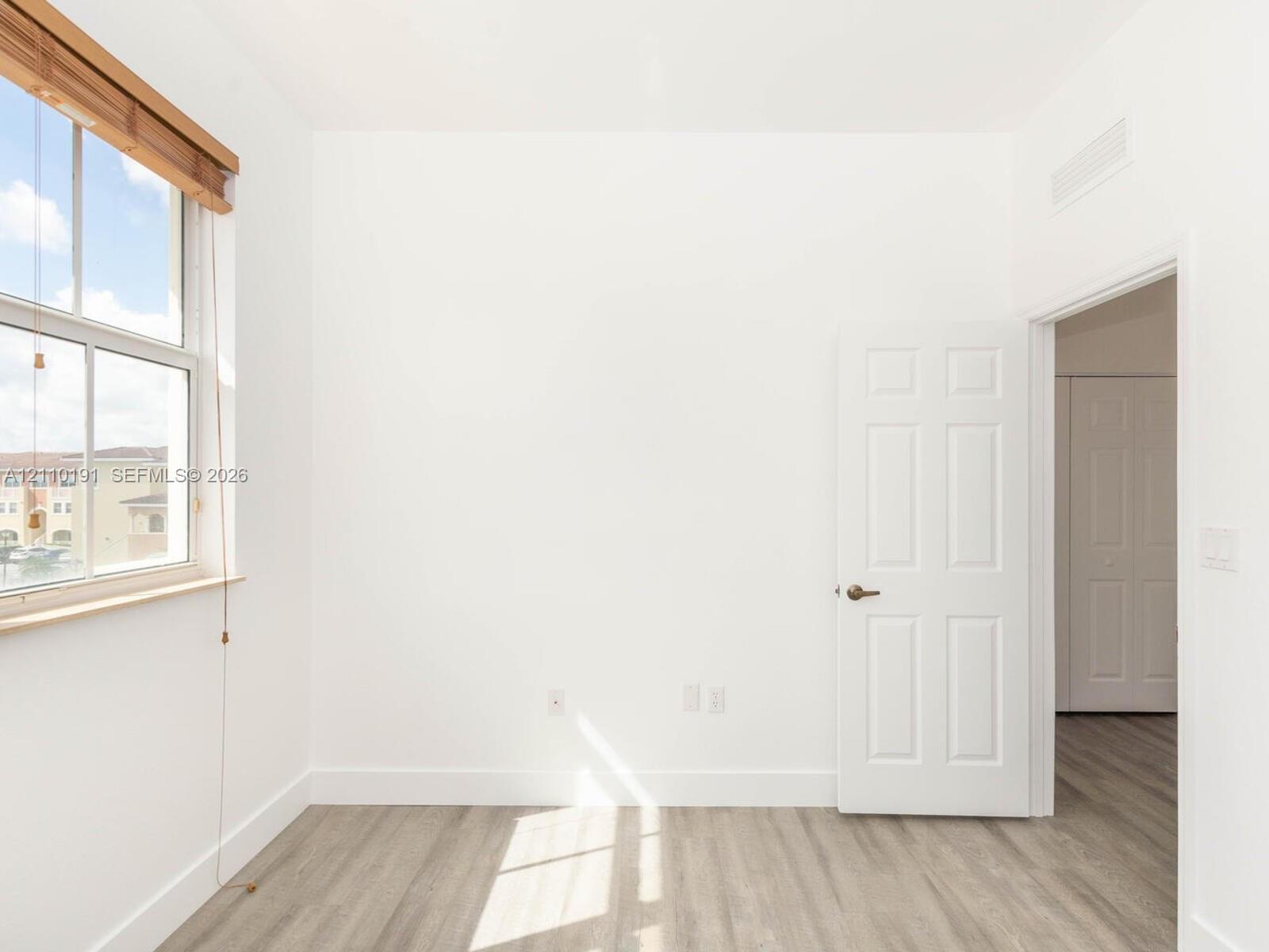Wood-finish flooring, multi-pane window with wood-trim framing, six-panel interior door, and smooth-finish walls